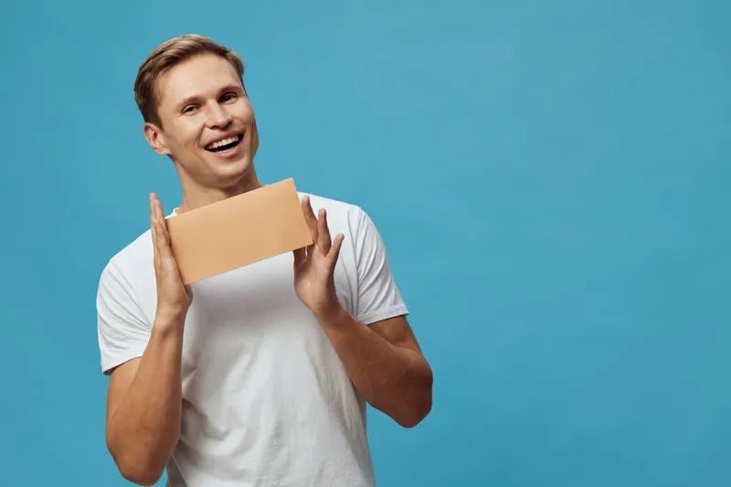 Smiling man holding blank cardboard sign with both hands, casual white t-shirt, blue background, positive emotions, studio portrait, happiness, advertising concept, people lifestyle.