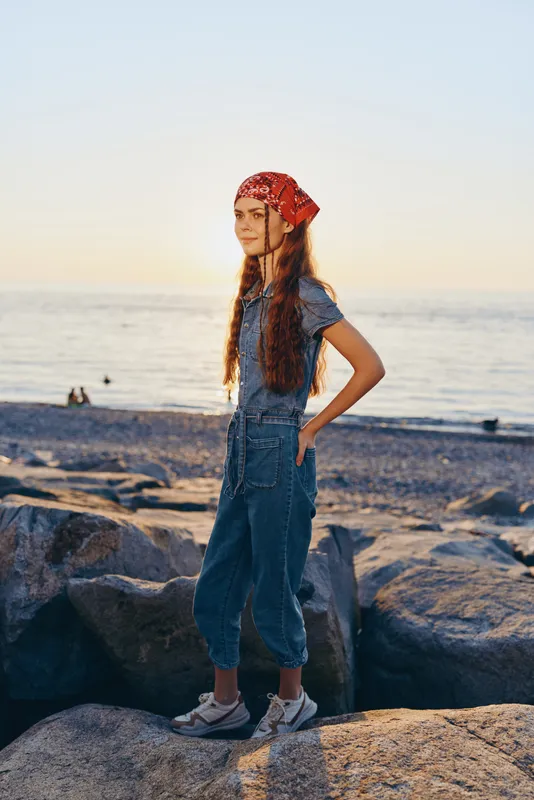 Lifestyle woman in modern boho-western denim street style stands on rocky beach at sunset, naturally smiling, film color tones enhancing warm atmosphere.