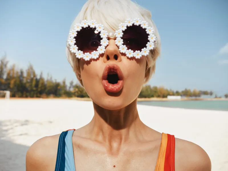 A stylish woman at a sunny beach wears whimsical round sunglasses made of daisies, with a surprised open-mouth expression, creating a bold, eye-catching fashion moment.