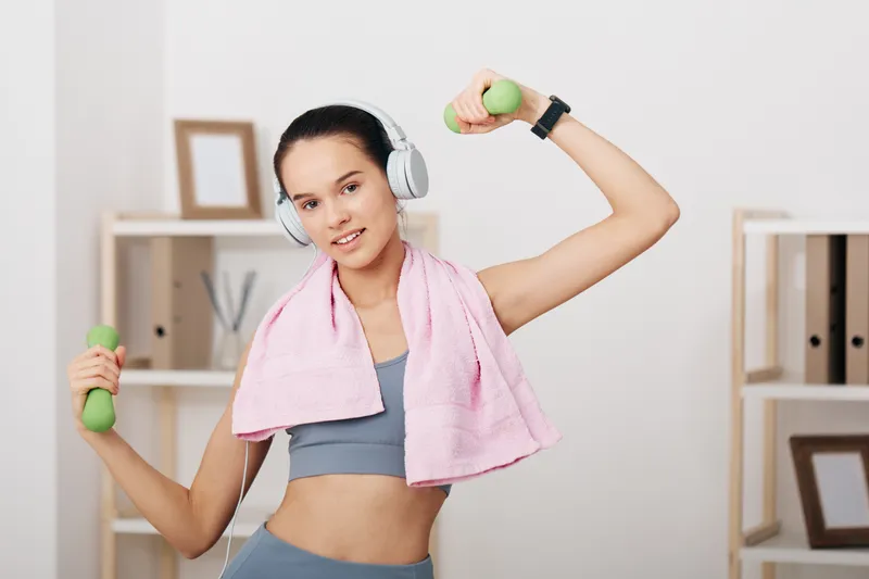 Fitness woman in a bright room lifts dumbbells with headphones on and a pink towel around her neck, presenting focused workout, strength training and home fitness.