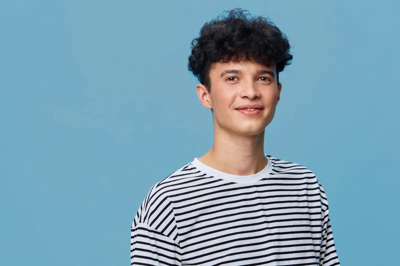 young man smiling wearing striped shirt standing against blue background, casual style, cheerful expression, studio portrait with soft lighting