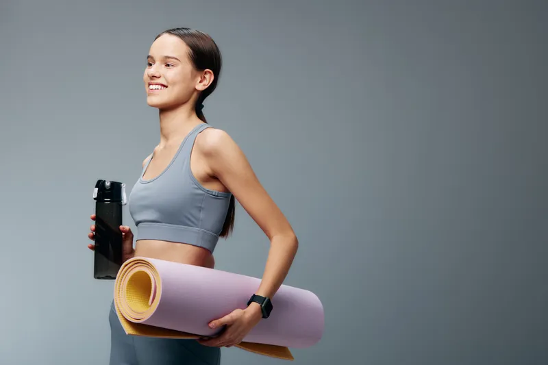 Woman in athletic wear holds a water bottle and rolled yoga mat, ready for a workout. Fitness, sport and yoga concept with studio vibe, motivation, balance and energy for a gym routine.