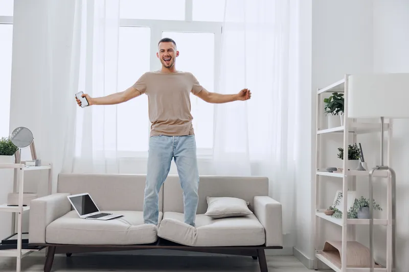 A joyful man standing on a couch, embracing a digital detox and showing soft masculinity in a bright living space, representing modern emotions and work-life blend.