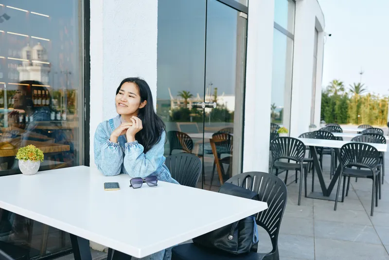 Woman sits at a white outdoor cafe table, hands near chin in thought, backpack and sunglasses beside, glass storefronts and cafe chairs in bright daytime ambiance, a calm moment