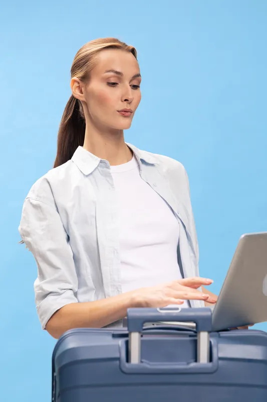 Young woman with suitcase and laptop looks focused on screen. Casual traveler prepares for trip wearing light shirt against blue background. Concept for travel, planning, and technology.