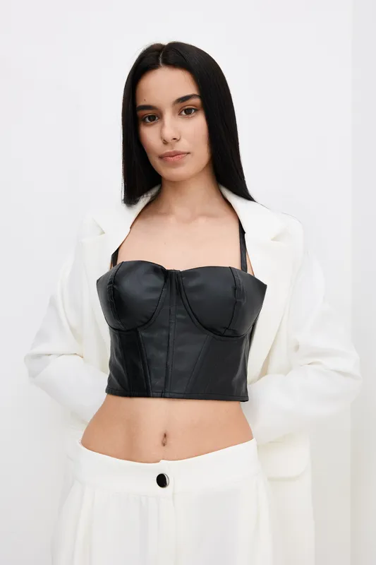 young woman wearing a black leather corset and white blazer standing against a plain white background with a confident and stylish pose