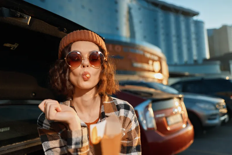 Woman sunglasses beanie golden hour candid urban food parking portrait playful fish-face holding takeaway in city lot, authenticity, candid lifestyle, golden hour glow, mindful living.