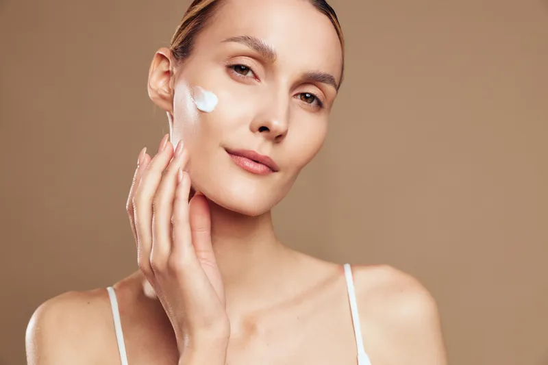 Young woman applies moisturizing cream on her face with fingers, shows natural skin care routine, smooth complexion, isolated on beige background, beauty concept.