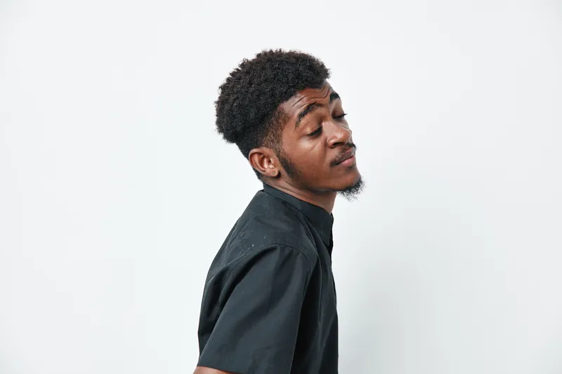 Young black man with dark skin and curly hair wearing black shirt posing with closed eyes and confident expression on white background in studio.