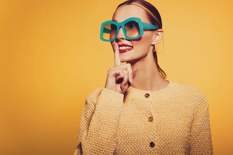 Stylish woman in yellow sweater and large blue sunglasses makes shushing gesture with finger on lips. Bright yellow background creates cheerful and playful mood for fashion or lifestyle concept.