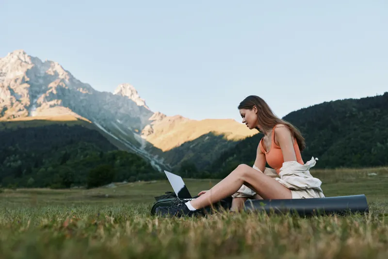 A solo woman sits on a yoga mat in a grassy field, using a laptop with mountains in the background. She wears active clothing and appears focused, enjoying a peaceful work break in nature.