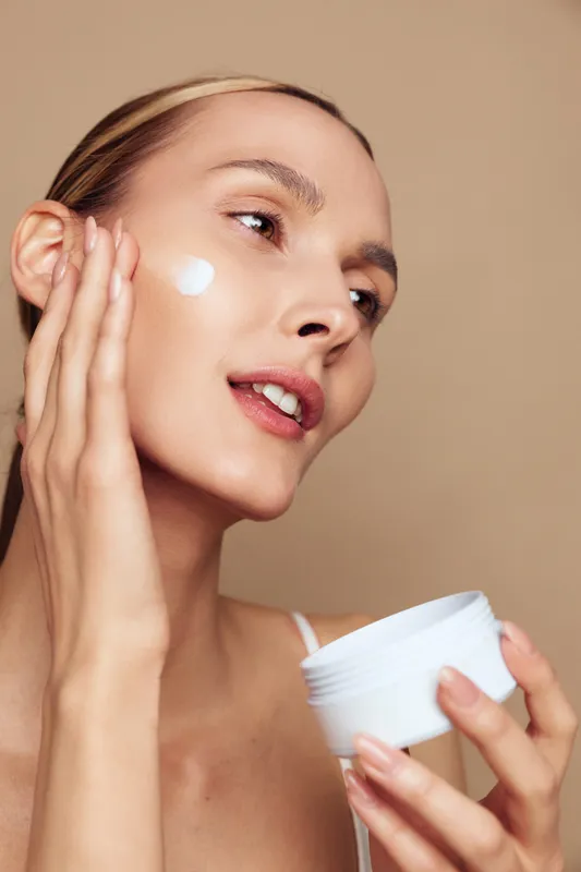 Young woman with clear skin applying white face cream on cheek. Close-up portrait on beige background. Concept of skincare, beauty routine, and natural wellness. Soft lighting highlights healthy skin.