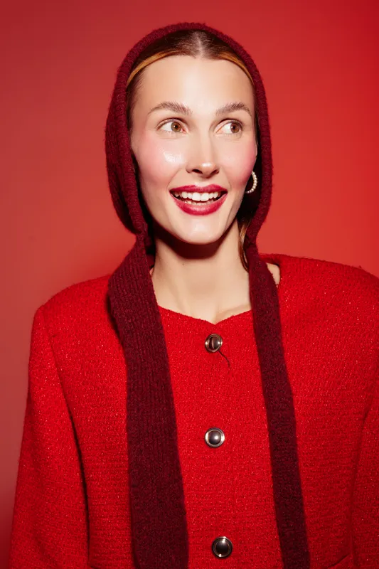 Smiling woman in red sweater and matching hood looks away with bright lipstick on red background. Concept of fashion, winter style, and cheerful mood in studio lighting.