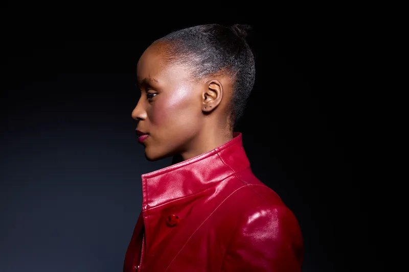Profile of an African woman with dark skin wearing a shiny red leather jacket against a black background. The image highlights fashion, beauty, and elegance in studio lighting.