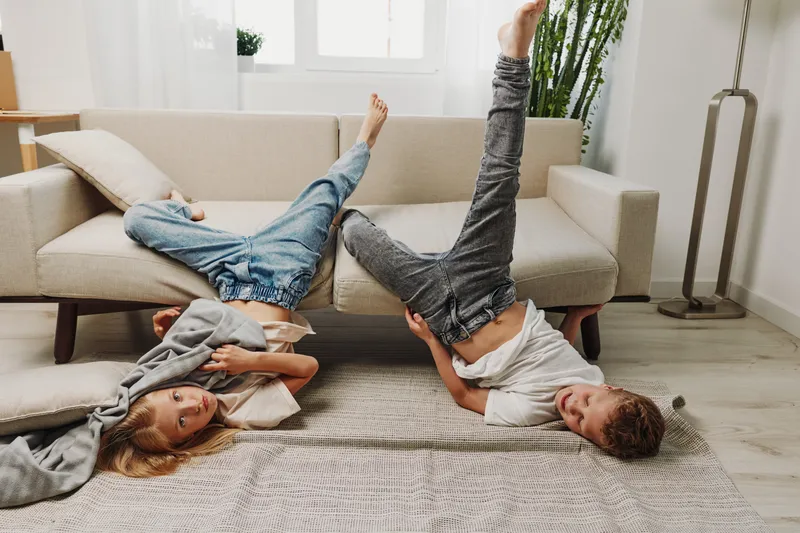 Two children in casual clothes playfully pose upside down on a rug near a beige sofa in a bright living room. The scene captures fun, childhood joy, and family leisure moments indoors.