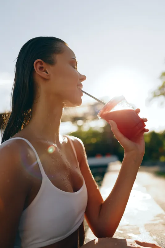Cheerful woman outdoors drinking a cool beverage through a straw, sunlit moment, relaxed posture, warm breeze, vibrant colors, poolside setting, summer lifestyle, candid and refreshing