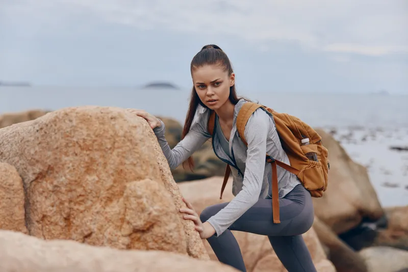 Young woman hiking on rocky beach, wearing athletic clothing and a backpack, displaying determination against a coastal background