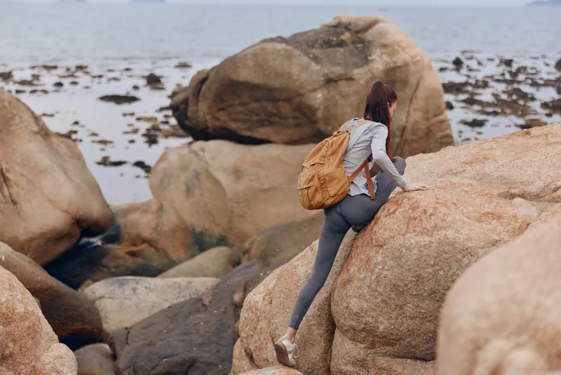 Young woman climbing rocky terrain near a calm sea, wearing athletic clothing and a brown backpack, expressing determination and a spirit of adventure