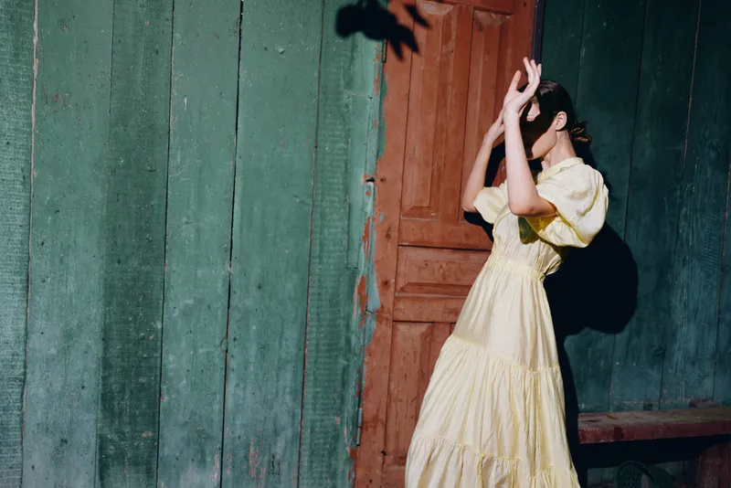 Woman in yellow dress shielding face from sunlight and shadow by rustic wooden door, vintage portrait with natural pose authenticity inclusivity in candid scene