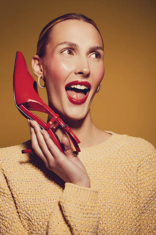 Happy woman with red lipstick holds red high heel shoe near face, smiling and looking aside. Fashion concept with bright colors and joyful expression on yellow background.