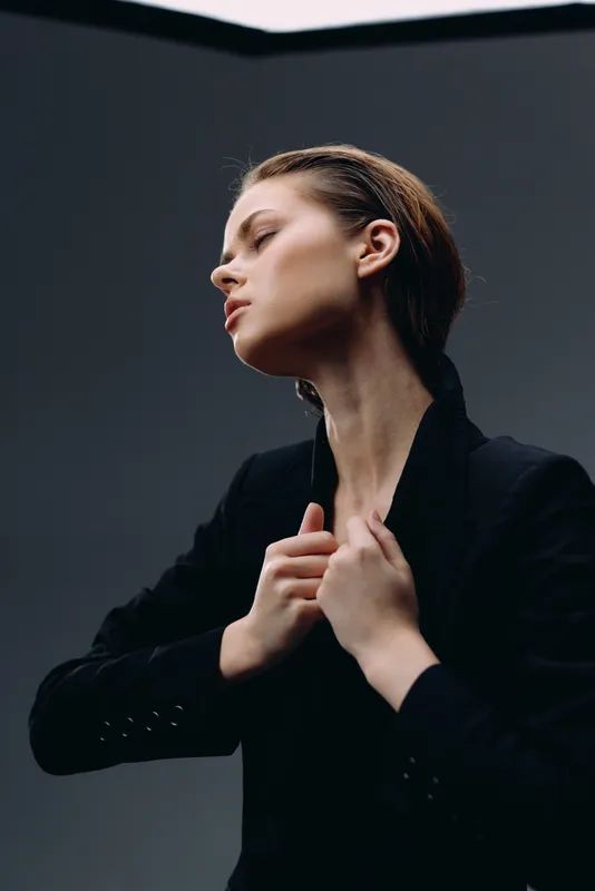 Elegant woman in black jacket posing with closed eyes, expressing calm and confidence on dark neutral background.