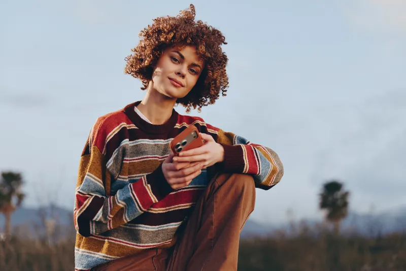 Smiling woman with curly hair outdoors holding a smartphone, casual lifestyle portrait in a striped sweater, warm light, blurred park background, daily communication