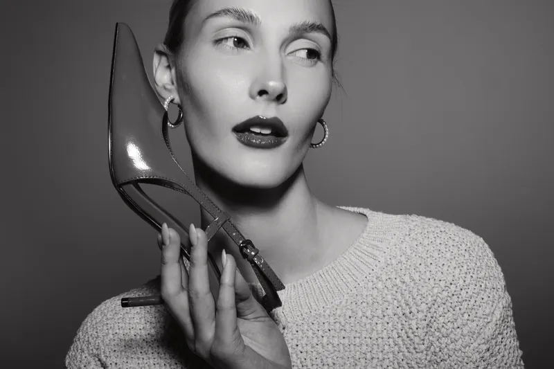 Young caucasian woman with pale skin holds high heel shoe near face, looking away. Black and white portrait with stylish expression and elegant makeup in studio lighting.
