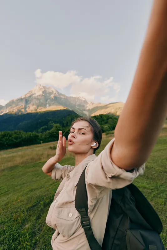 Young woman lifestyle traveler takes selfie on mountain trail with backpack wears wireless earbuds enjoys outdoor adventure in nature landscape under clear sky.