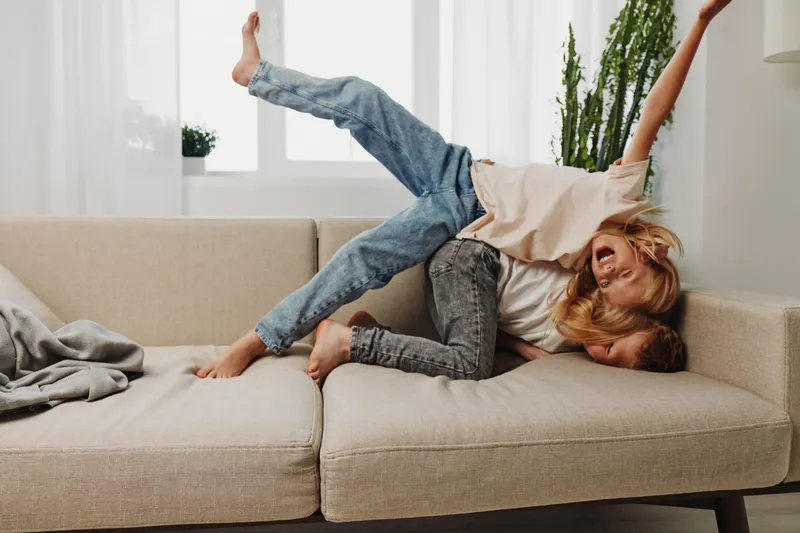 Happy playful children wrestling on beige sofa in bright living room with plants, joyful family moment and fun at home concept.