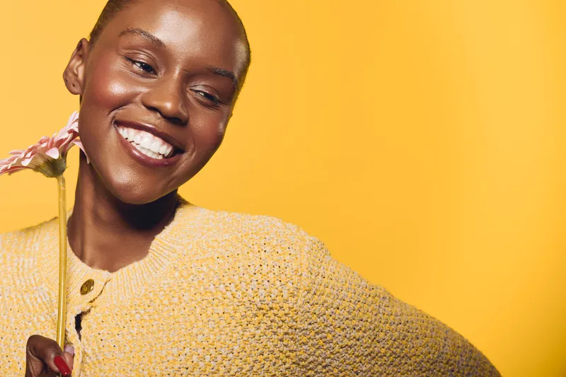 Joyful black woman with dark skin wearing yellow sweater smiles holding pink flower on yellow background. Concept of happiness, beauty, and natural glow in studio portrait.