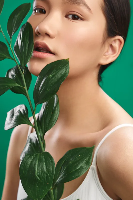 asian woman with smooth complexion poses beside large green leaves against a vibrant green backdrop, showcasing natural beauty and calm confidence in a studio portrait.