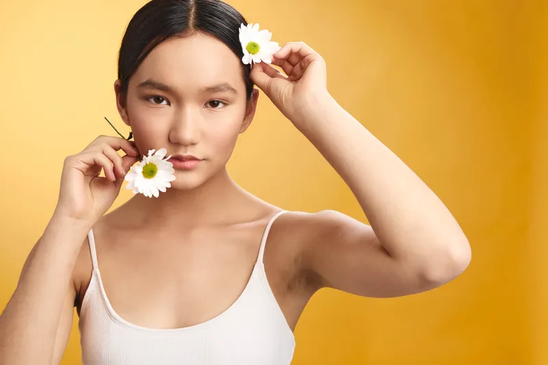 Asian woman holds white daisies near her face against a bright yellow backdrop. She wears a white tank top and gazes softly at the camera, forming a fresh, artistic portrait mood.