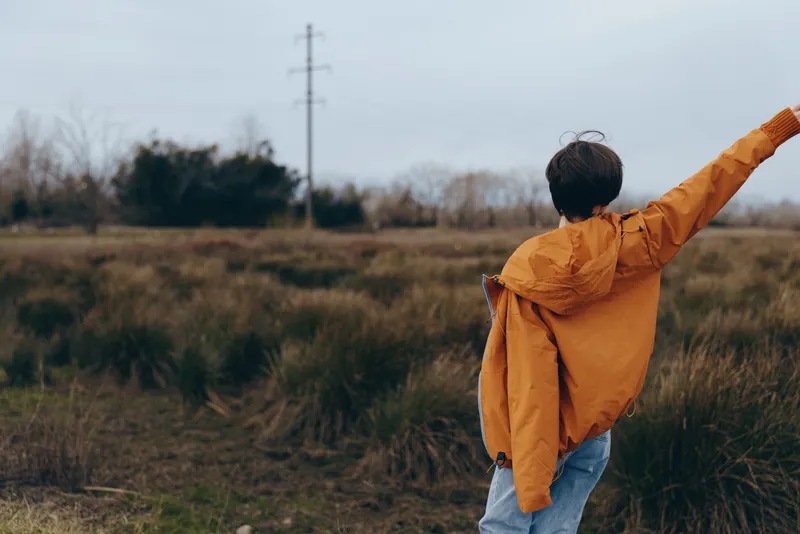 Woman outdoors in a grassy field, wearing an orange jacket, lifting her arms in a joyful lifestyle moment, soft wind, natural light, blurred trees background