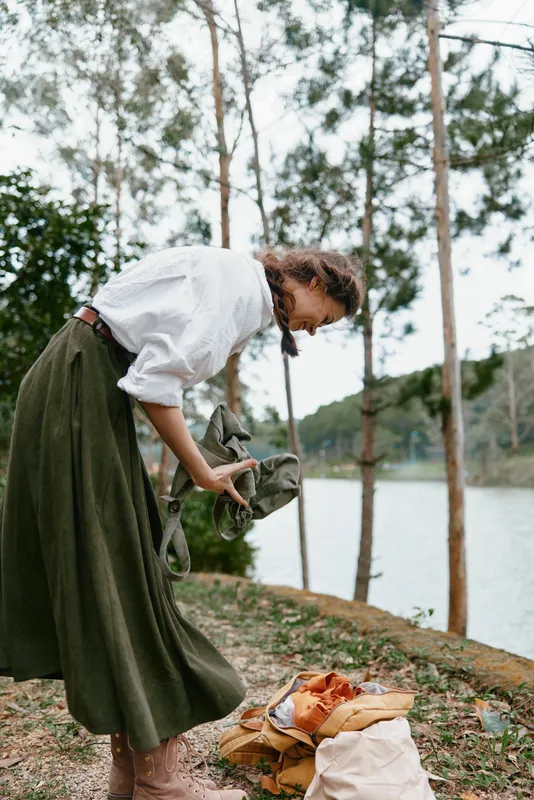 Young woman in nature with green skirt and white blouse, joyfully organizing autumn leaves near a river, evoking a sense of connection to nature and seasonal change