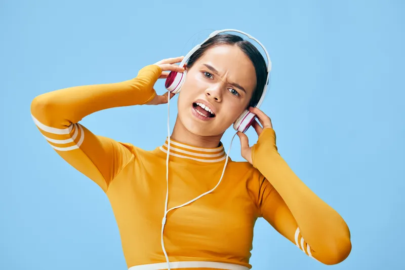 Young athletic woman in a yellow fitness top wearing headphones, energetic workout pose and focused expression against a soft blue studio background, sport music motivation.