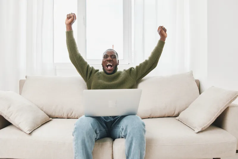 Excited African man celebrating success while working on a laptop at home in a bright, cozy living room during a sunny day