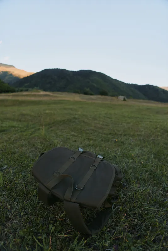 A lone backpack rests on a grassy field with distant rolling hills under a clear blue sky, conveying solitude and a quiet mood of outdoor travel and exploration.