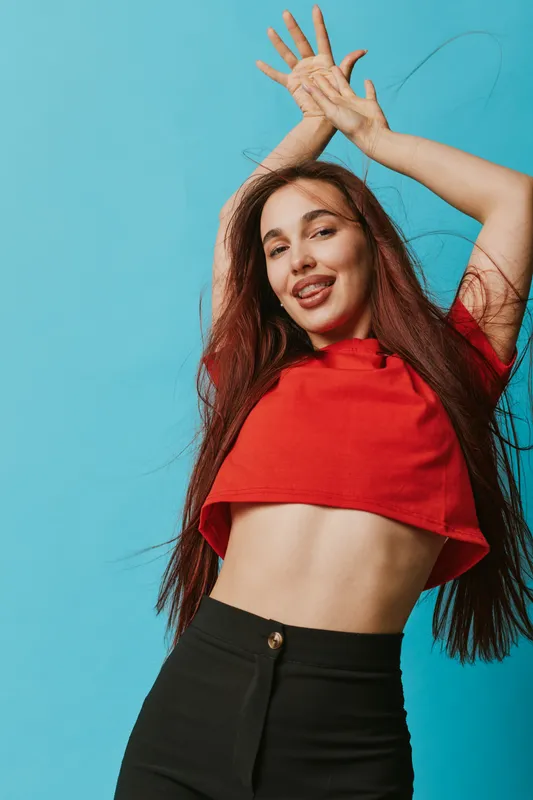 Young woman with long hair wearing red crop top and black pants posing with hands raised and playful expression against blue background in studio. Casual style and confident mood.