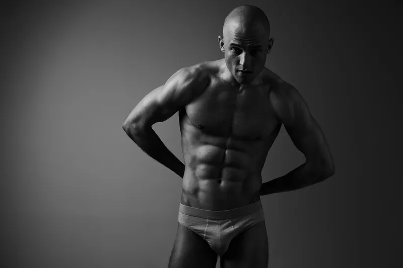 Black and white image of a muscular man posing confidently, showcasing his toned physique and athleticism in a dramatic light setting.