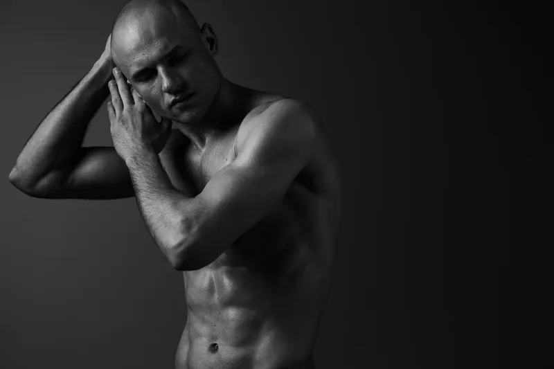 A contemplative man poses in black and white, portraying vulnerability and strength, showcasing emotional depth in a minimalistic setting.