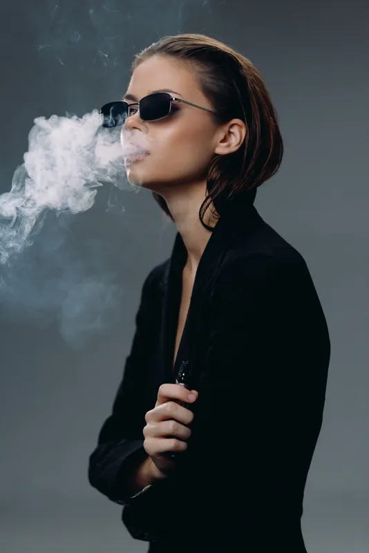 Woman wearing sunglasses blowing smoke with confident expression in dark stylish outfit