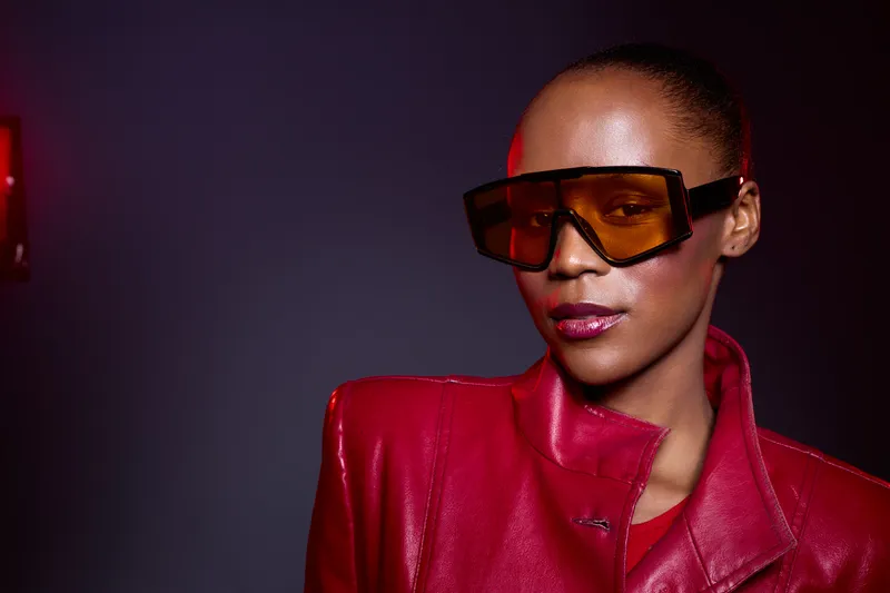 Confident black woman with dark skin wearing oversized orange sunglasses and a red leather jacket poses against a dark background. Fashion, style, and modern portrait concept.