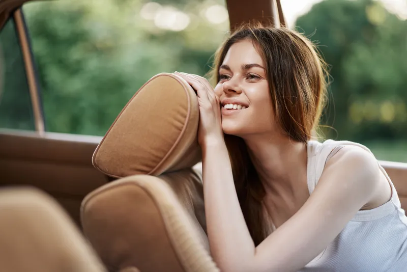 Young woman in casual attire smiling while sitting in a car, enjoying a sunny day, showcasing joy and relaxation in a natural outdoor setting.