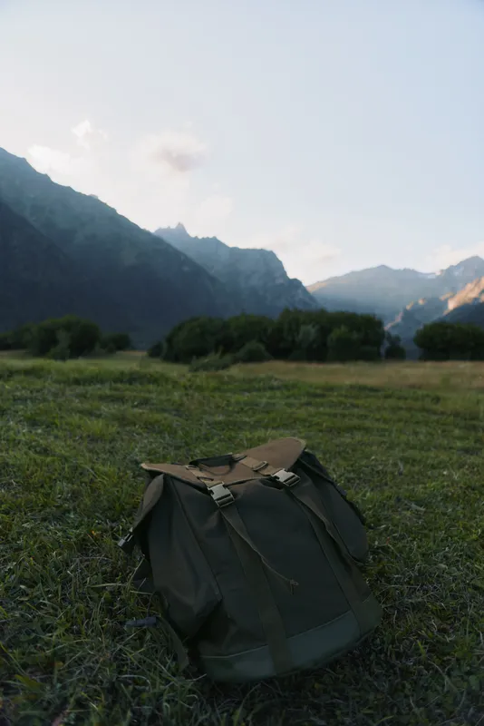 Backpack rests in a grassy field with distant mountains, suggesting a solo travel mood, outdoor adventure, and calm nature moment. The silent scene invites exploration, preparation for a hike, and