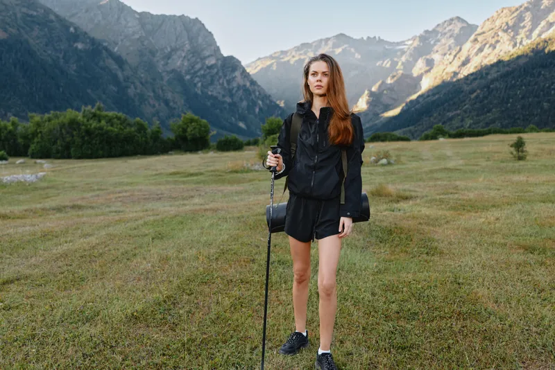 A lone woman hiking in a rugged mountain valley. She stands with a trekking pole, wearing a black jacket and shorts, looking ahead with calm confidence in a vast outdoor landscape.