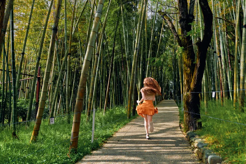 Girl running on a bamboo forest path in a flowing orange dress, back view and motion capture, sunlight filtering through tall trees, nature trail with peaceful summer scenery