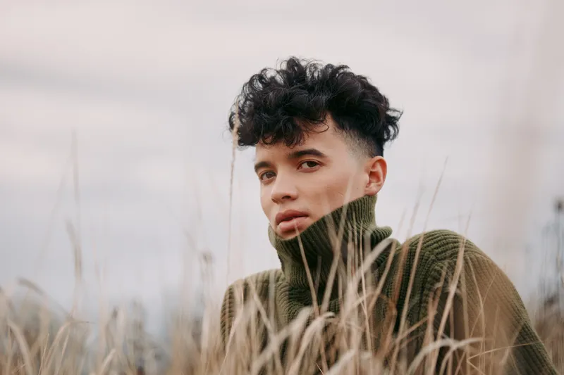 Candid portrait of a young man in a warm turtleneck among tall dry grasses, natural gaze and subtle emotion conveying a sense of genuine presence and honest personal expression.
