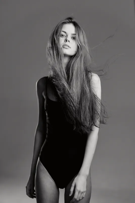 Black and white portrait of a slender woman with long hair wearing a black bodysuit on a gray background, expressing calm and confidence.