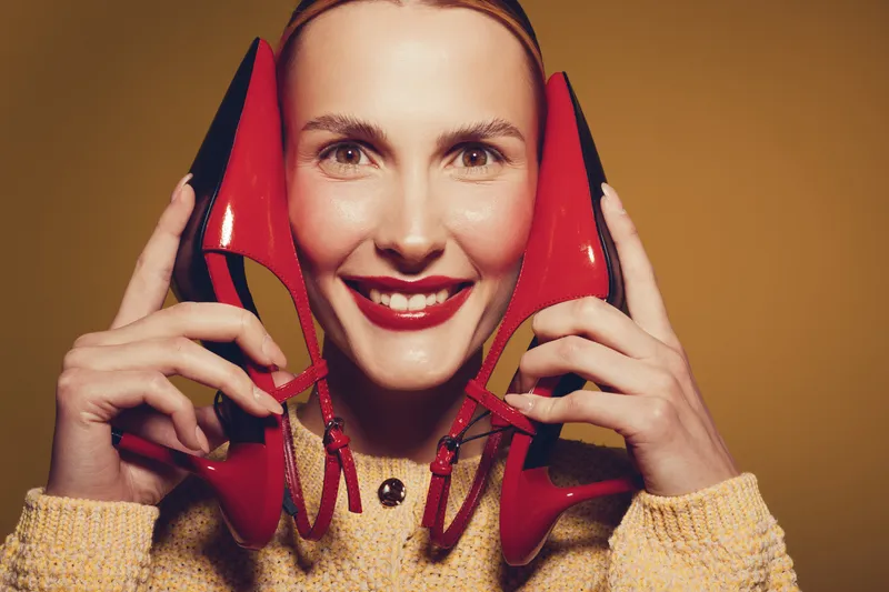 Smiling woman holds red high heels near face, wearing yellow sweater. Studio portrait with warm lighting on brown background. Concept of fashion, style, and happiness.