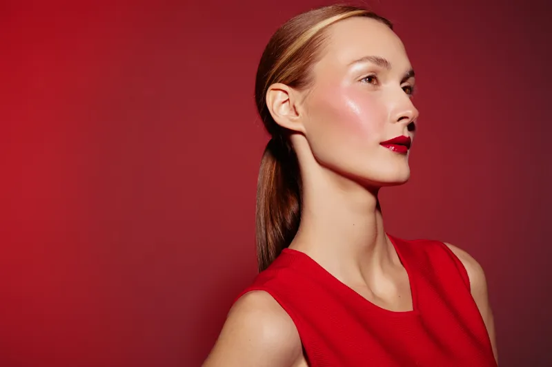 Young woman with shiny skin and red lipstick poses in red sleeveless top against red background. Concept of beauty, fashion, makeup, and confident female portrait.